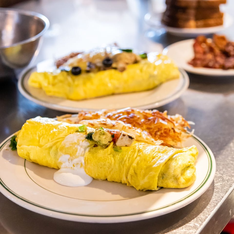 Golden omelets filled with meat and vegetables sit on plates at a restaurant counter, with pancakes and bacon in the background.
